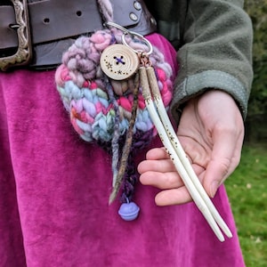 May include: A pink crocheted pouch with a wooden button and two wooden sticks hanging from it. The pouch is attached to a brown leather belt with a metal buckle.