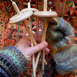 Puede incluir: Un conjunto de husos de madera con acabado natural, sostenidos en una mano. Los husos tienen un disco circular y un gancho en la parte superior. Tambi&eacute;n se ve lana colorida en tonos verdes, azules y morados. El fondo es un textil estampado.