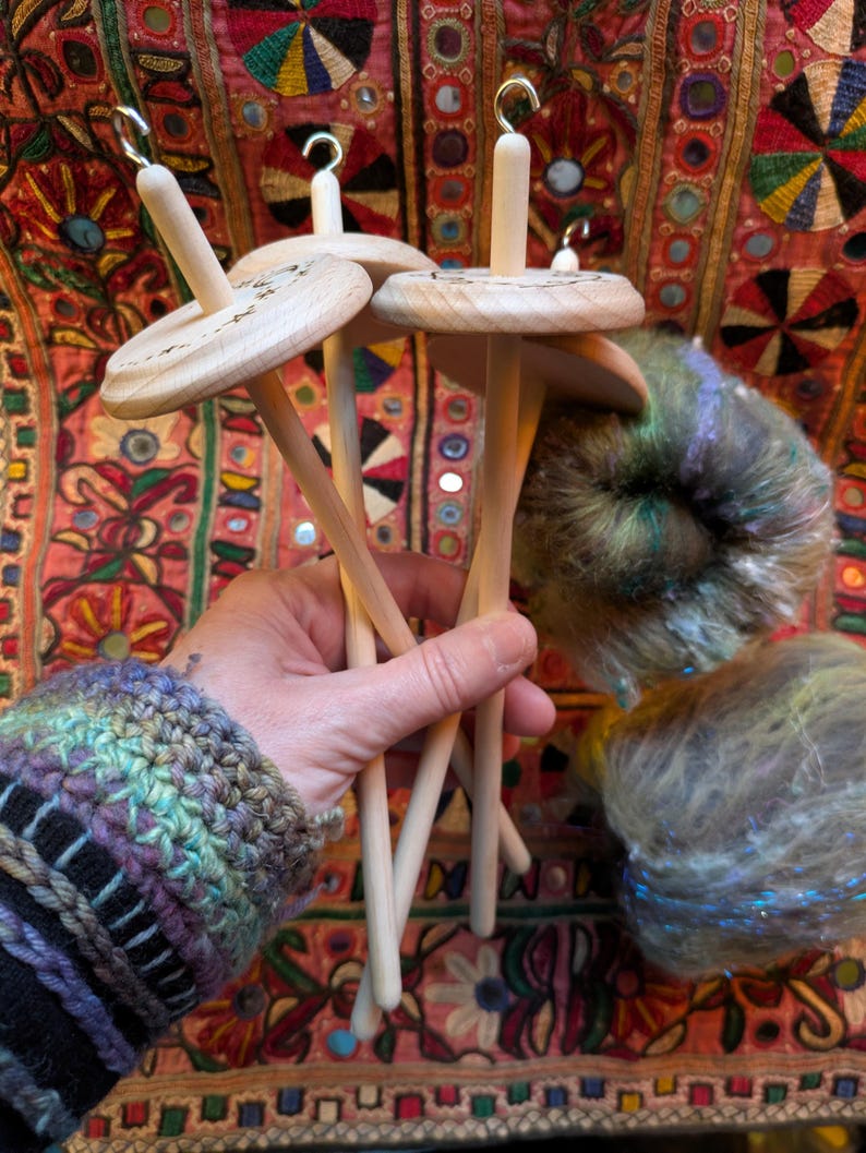 Puede incluir: Un conjunto de husos de madera con acabado natural, sostenidos en una mano. Los husos tienen un disco circular y un gancho en la parte superior. Tambi&eacute;n se ve lana colorida en tonos verdes, azules y morados. El fondo es un textil estampado.