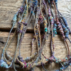 May include: A collection of handmade fiber art pieces, featuring strands of yarn in various colors. The yarns are primarily in shades of blue, purple, and gold, with textured details. The pieces are arranged against a rustic wooden background.
