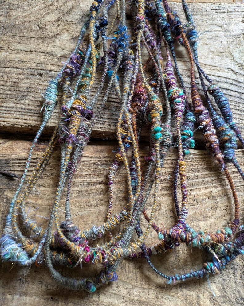 May include: A collection of handmade fiber art pieces, featuring strands of yarn in various colors. The yarns are primarily in shades of blue, purple, and gold, with textured details. The pieces are arranged against a rustic wooden background.