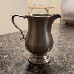 May include: A small, antique pewter creamer with a heart-shaped handle and a spout. The creamer has a bulbous body and sits on a round base. The pewter has a dull, metallic finish.