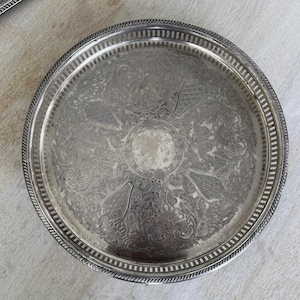 May include: A round, silver-colored tray with an ornate, engraved floral design. The tray has a detailed border and a rope-like edge. Another tray is partially visible in the background.