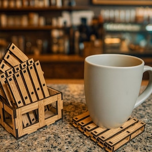 May include: A white ceramic mug sits on a wooden pallet-style coaster. Several similar coasters are stacked in a matching wooden box. The coasters and box are made of light-coloured wood with dark brown accents.