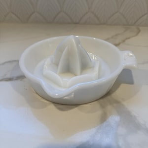 May include: A white ceramic citrus juicer with a reamer and a small handle. The juicer has a round bowl and a ridged cone-shaped reamer in the center. The kitchen tool is designed for extracting juice from citrus fruits.