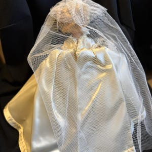 Puede incluir: Una mu&ntilde;eca vintage vestida con un vestido de novia de sat&eacute;n y un velo de tul. El vestido es de color marfil cremoso y el velo es blanco. La mu&ntilde;eca est&aacute; sobre un fondo oscuro, creando un contraste.