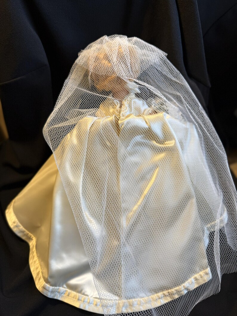 Puede incluir: Una mu&ntilde;eca vintage vestida con un vestido de novia de sat&eacute;n y un velo de tul. El vestido es de color marfil cremoso y el velo es blanco. La mu&ntilde;eca est&aacute; sobre un fondo oscuro, creando un contraste.