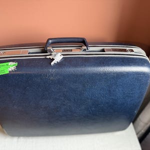 May include: A dark blue hard-shell suitcase with a textured finish, a dark blue top handle, and silver-toned metal latches. A small silver key hangs from the handle. A green sticker on the left side reads British Airways Security Screened.