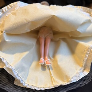 Puede incluir: Una mu&ntilde;eca vintage con un vestido color crema con ribete de encaje blanco y una enagua de tul. Se ven las piernas y los pies de la mu&ntilde;eca, con sandalias blancas. El vestido est&aacute; levantado, revelando la ropa interior de la mu&ntilde;eca.
