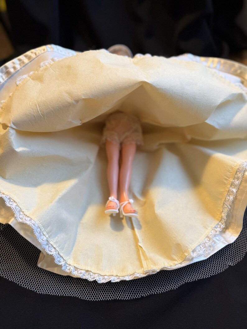 Puede incluir: Una mu&ntilde;eca vintage con un vestido color crema con ribete de encaje blanco y una enagua de tul. Se ven las piernas y los pies de la mu&ntilde;eca, con sandalias blancas. El vestido est&aacute; levantado, revelando la ropa interior de la mu&ntilde;eca.