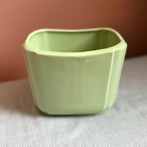 May include: A light green ceramic planter with a square shape and rounded corners. The vessel features vertical indentations along each side, creating a subtle paneled effect. The interior is hollow and the exterior has a smooth, matte finish.