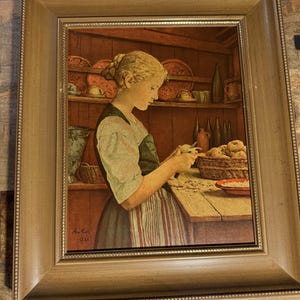May include: Framed print of a woman peeling potatoes in a kitchen. The artwork features a woman in a light green and dark blue dress, with a red and white striped apron. The print is signed "Anker 1886" in the lower left corner.