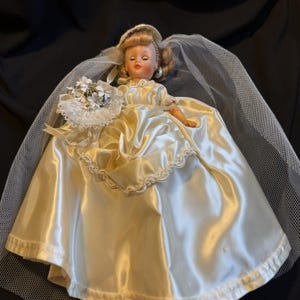 Puede incluir: Una mu&ntilde;eca vintage vestida de novia, con un vestido de novia de sat&eacute;n y un velo. La mu&ntilde;eca tiene el pelo rubio y sostiene un peque&ntilde;o ramo de flores blancas. El vestido tiene un adorno decorativo.