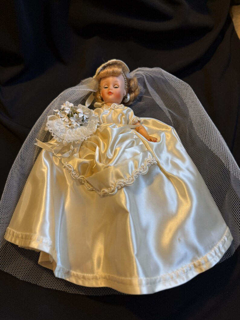 Puede incluir: Una mu&ntilde;eca vintage vestida de novia, con un vestido de novia de sat&eacute;n y un velo. La mu&ntilde;eca tiene el pelo rubio y sostiene un peque&ntilde;o ramo de flores blancas. El vestido tiene un adorno decorativo.