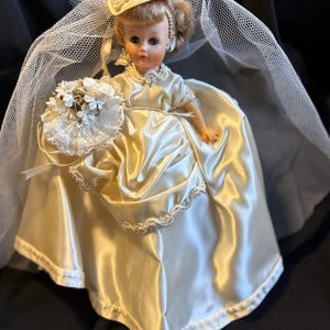 Puede incluir: Mu&ntilde;eca vintage vestida con un vestido de novia de sat&eacute;n, sombrero y velo. La mu&ntilde;eca sostiene un ramo de flores blancas. El vestido tiene un ribete festoneado. La mu&ntilde;eca tiene el pelo rubio y lleva pendientes.