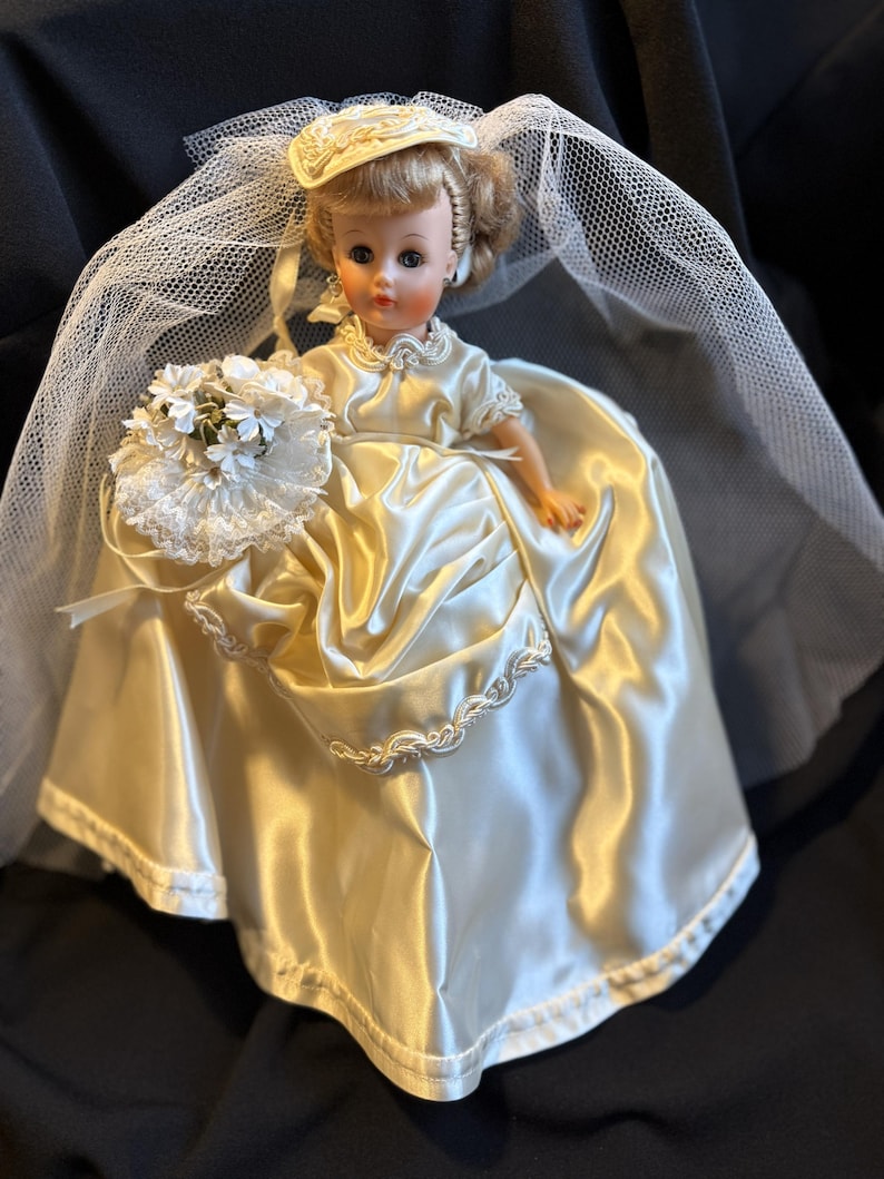 Puede incluir: Mu&ntilde;eca vintage vestida con un vestido de novia de sat&eacute;n, sombrero y velo. La mu&ntilde;eca sostiene un ramo de flores blancas. El vestido tiene un ribete festoneado. La mu&ntilde;eca tiene el pelo rubio y lleva pendientes.