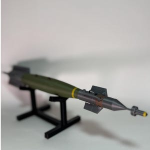 Peut inclure: Maquette d'une bombe militaire, peinte en vert olive avec des détails gris et jaunes. La bombe est présentée sur un support noir sur fond blanc. Le modèle a un corps cylindrique et un nez pointu.