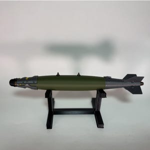 Puede incluir: Modelo de bomba en verde oliva y gris metálico, exhibido sobre un soporte negro. La bomba presenta una nariz detallada, aletas y puntas de estabilización. El diseño sugiere un tema militar o histórico, con una longitud aproximada de 70 cm.