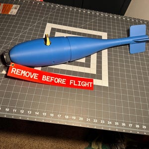 Op de afbeelding: Een blauw model van een bom met een vinstabilisator en een rood label met de tekst "REMOVE BEFORE FLIGHT". Het model heeft een cilindrisch lichaam en een zwarte dop aan één uiteinde. Een goudkleurige pin is in het lichaam gestoken.
