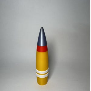 Puede incluir: Objeto cilíndrico con una punta plateada, una banda roja y un cuerpo amarillo. Dos bandas blancas rodean la parte inferior. El objeto está de pie sobre un fondo liso. El diseño es llamativo.