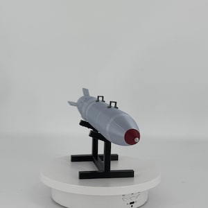 Op de afbeelding: Een grijs model van een bom met een rode neuskegel en zwarte handvatten, gemonteerd op een zwarte standaard. Het model wordt tentoongesteld op een wit draaiplateau tegen een witte achtergrond.