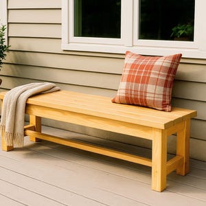 May include: A wooden bench with a light finish, a plaid pillow, and a folded beige throw. The bench is on a deck with two potted plants. The pillow has a red and white plaid pattern. The plants are in terracotta pots.