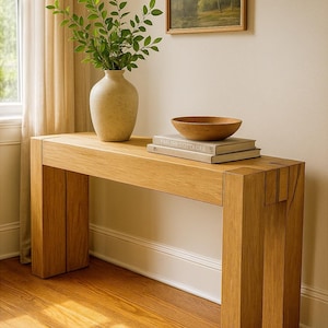 以下が含まれることがあります： 長方形の天板と4本の太い脚を持つ、明るいオーク材のコンソールテーブル。緑の葉が入ったベージュのセラミック花瓶、木製のボウル、本がテーブルの上にあります。額入りの風景画が壁に掛けられています。