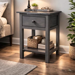 May include: A gray wooden nightstand with a drawer and a lower shelf. The nightstand has a dark knob and is holding a woven basket, books, and decorative items. The top of the nightstand has a lamp, a small plant, and glasses. The text "DIGITAL DOWNLOAD WOODWORKING PLANS" is visible.