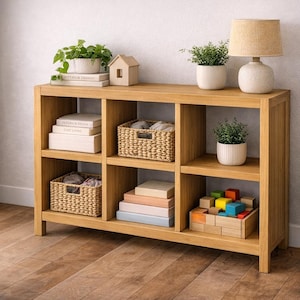 Puede incluir: Un mueble de almacenamiento de madera clara con seis estantes cúbicos. Los estantes contienen libros, cestas tejidas, plantas en macetas, una lámpara y bloques de madera. La unidad se coloca contra una pared clara y un suelo de madera.
