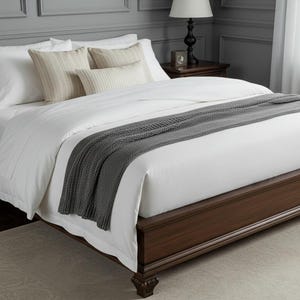 Puede incluir: Una cama con ropa de cama blanca, almohadas y una manta de punto gris. El marco de la cama, de color marrón oscuro, tiene patas talladas. La habitación tiene paredes grises, una alfombra y una mesita de noche con una lámpara.