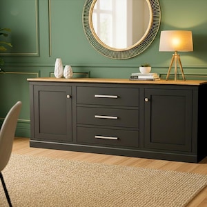 May include: A dark grey sideboard with three drawers and two cabinet doors, topped with a light wood surface. The sideboard is in a room with a green wall, a round mirror, and a beige rug.