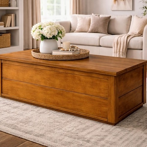 May include: A rectangular wooden coffee table with a warm, medium-brown finish. The table has a simple, modern design with two visible drawers. A woven tray with a white vase of flowers and a candle sits on top. The table is in a living room setting.