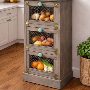 May include: A wooden kitchen storage cabinet with three wire mesh drawers. The cabinet is gray with brass accents and turquoise knobs. The drawers contain various vegetables, including potatoes, onions, and oranges. The cabinet is on a wooden floor.