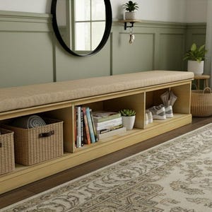 以下が含まれることがあります： ベージュのクッションのトップとオープン収納コンパートメントを備えた明るい木製のベンチ。2つの織りバスケットには、左側のコンパートメントに丸められたタオルが入っています。本、植物、靴は他のコンパートメントに見えます。装飾的なラグが前景にあります。