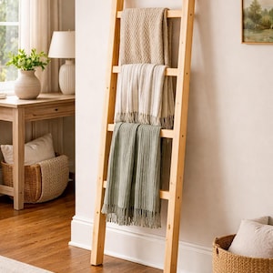 May include: A wooden blanket ladder displays three folded throws in beige, cream, and sage green. The room has a neutral colour palette with a wooden floor, a woven basket, and a framed landscape painting. The ladder is against a wall.