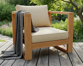 May include: A wooden outdoor armchair with a beige cushion and pillow. A gray blanket is draped over the arm. The chair is on a wooden deck, with a garden in the background.