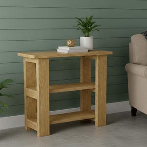 Puede incluir: Una mesa auxiliar de madera de roble claro con dos estantes. La mesa es rectangular con un diseño simple y robusto. Una pequeña planta en maceta y objetos decorativos están encima. La mesa está colocada frente a una pared verde, junto a un sillón beige.