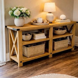 Könnte beinhalten: Eine helle Holzkonsolentisch mit zwei Regalen und X-förmigen Stützen. Auf der oberen Ablage stehen eine Vase mit weißen Blumen, eine Lampe und dekorative Gegenstände. Geflochtene Körbe und Bücher befinden sich auf den unteren Regalen.