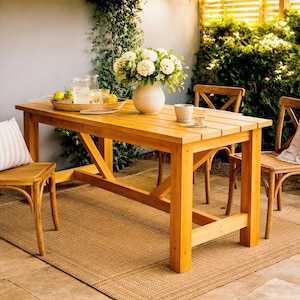 May include: A wooden garden dining set featuring a rectangular table and four chairs. The table, made of light-coloured wood, has a cross-leg design. A vase of white flowers sits on the table, alongside a tray of drinks. The chairs have curved backs.