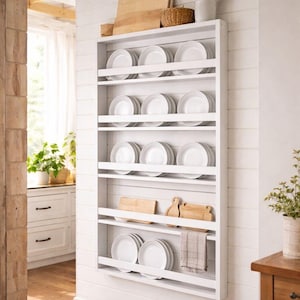May include: A white, wall-mounted plate rack filled with white plates. The rack has multiple shelves and is adorned with wooden cutting boards, a small basket, and utensils. A neutral-toned kitchen setting with wooden floors and white walls.