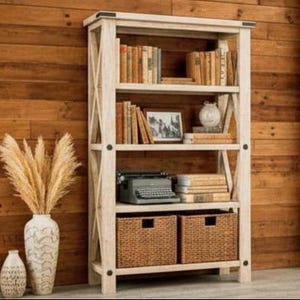May include: A rustic, off-white wooden bookshelf with multiple shelves filled with books, a framed photo, and decorative items. Two woven baskets sit at the bottom. A vintage typewriter is on one of the shelves. The bookshelf is set against a wooden wall.