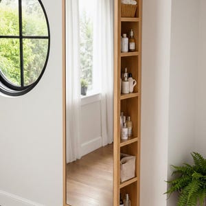 May include: A full-length wooden mirror with integrated shelving. The mirror is framed in light wood and has open shelves on the right, holding toiletries and a basket. A round window is visible on the left.