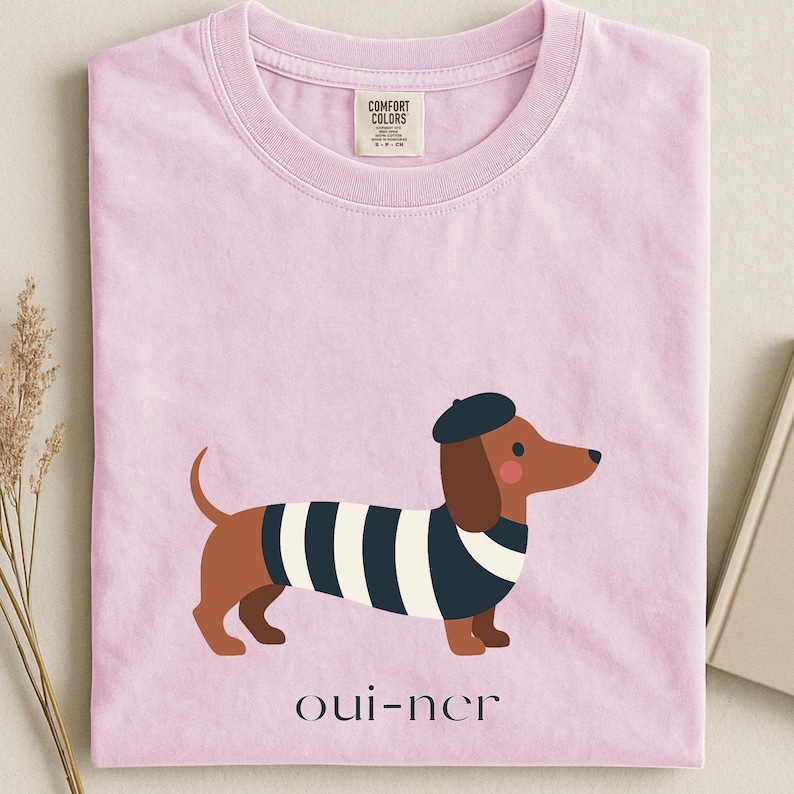 May include: A light pink t-shirt featuring a cartoon dachshund wearing a beret and a striped shirt. The text "oui-ner" is printed below the dog. The shirt has a Comfort Colors tag.