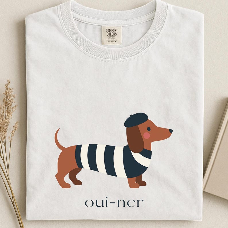 May include: White t-shirt featuring a cartoon dachshund wearing a beret and a striped shirt. The dog is brown with black and white stripes. The text "oui-ner" is printed below the dog.