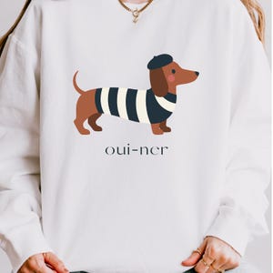 May include: White sweatshirt featuring a cartoon dachshund wearing a beret and a striped shirt. The dachshund is brown and black, with a red nose. The text "oui-ner" is printed below the dog in a simple font.