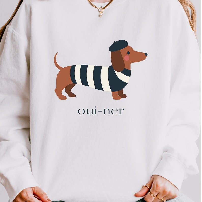 May include: White sweatshirt featuring a cartoon dachshund wearing a beret and a striped shirt. The dachshund is brown and black, with a red nose. The text "oui-ner" is printed below the dog in a simple font.