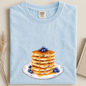 May include: Light blue t-shirt featuring a stack of pancakes with blueberries and syrup on a decorative plate. The pancakes are golden brown, and the blueberries are a deep blue. The shirt has a Comfort Colors tag.