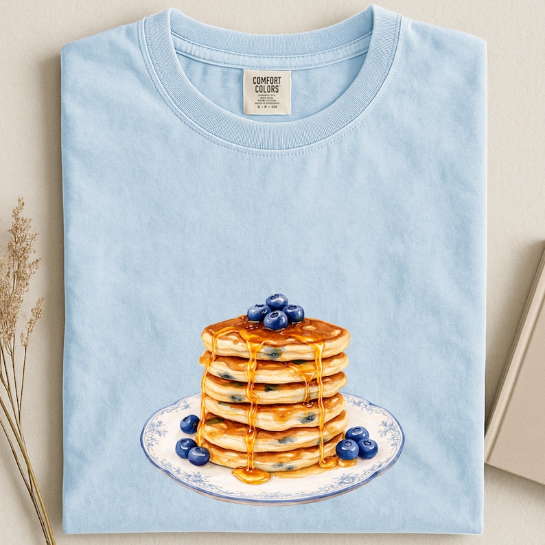 May include: Light blue t-shirt featuring a stack of pancakes with blueberries and syrup on a decorative plate. The pancakes are golden brown, and the blueberries are a deep blue. The shirt has a Comfort Colors tag.