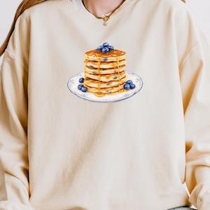 May include: Cream-colored sweatshirt featuring a graphic of a stack of pancakes with blueberries and syrup on a blue and white plate. The pancakes are golden brown, and the blueberries are a deep blue. The syrup is dripping down the sides.