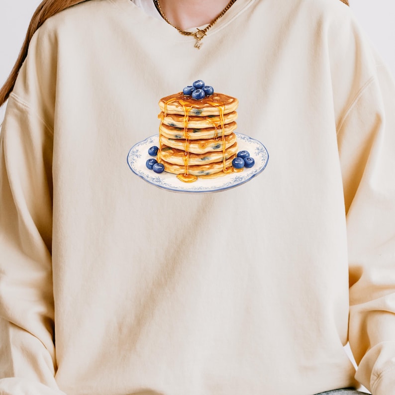 May include: Cream-colored sweatshirt featuring a graphic of a stack of pancakes with blueberries and syrup on a blue and white plate. The pancakes are golden brown, and the blueberries are a deep blue. The syrup is dripping down the sides.